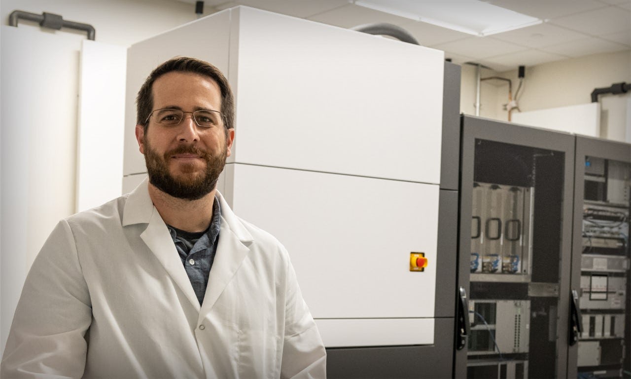 Jason McLellan wearing a white lab coat standing in front of the cryo-electron microscope at The University of Texas at Austin. 