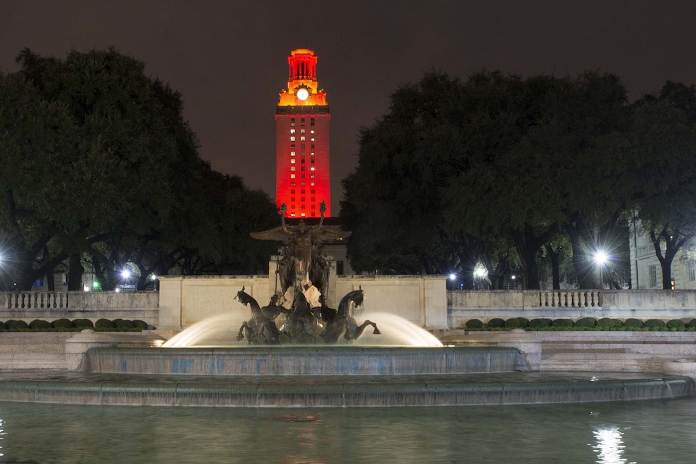 Night photo of the Little Field Fountain, a water feature with rearing horses in center of a round body of water the foreground and in the background the UT Tower lit with Orange lights