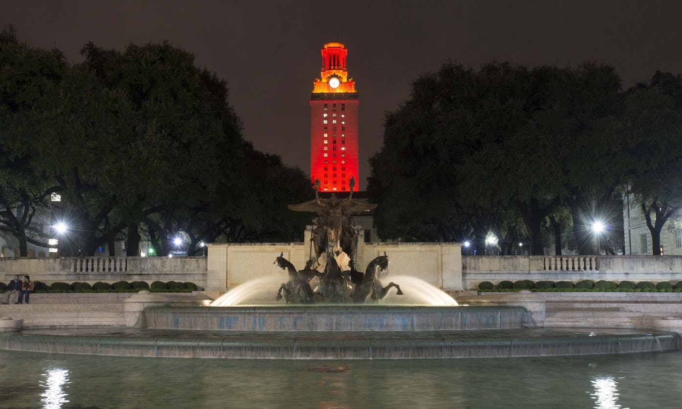 Night photo of the Little Field Fountain, a water feature with rearing horses in center of a round body of water the foreground and in the background the UT Tower lit with Orange lights